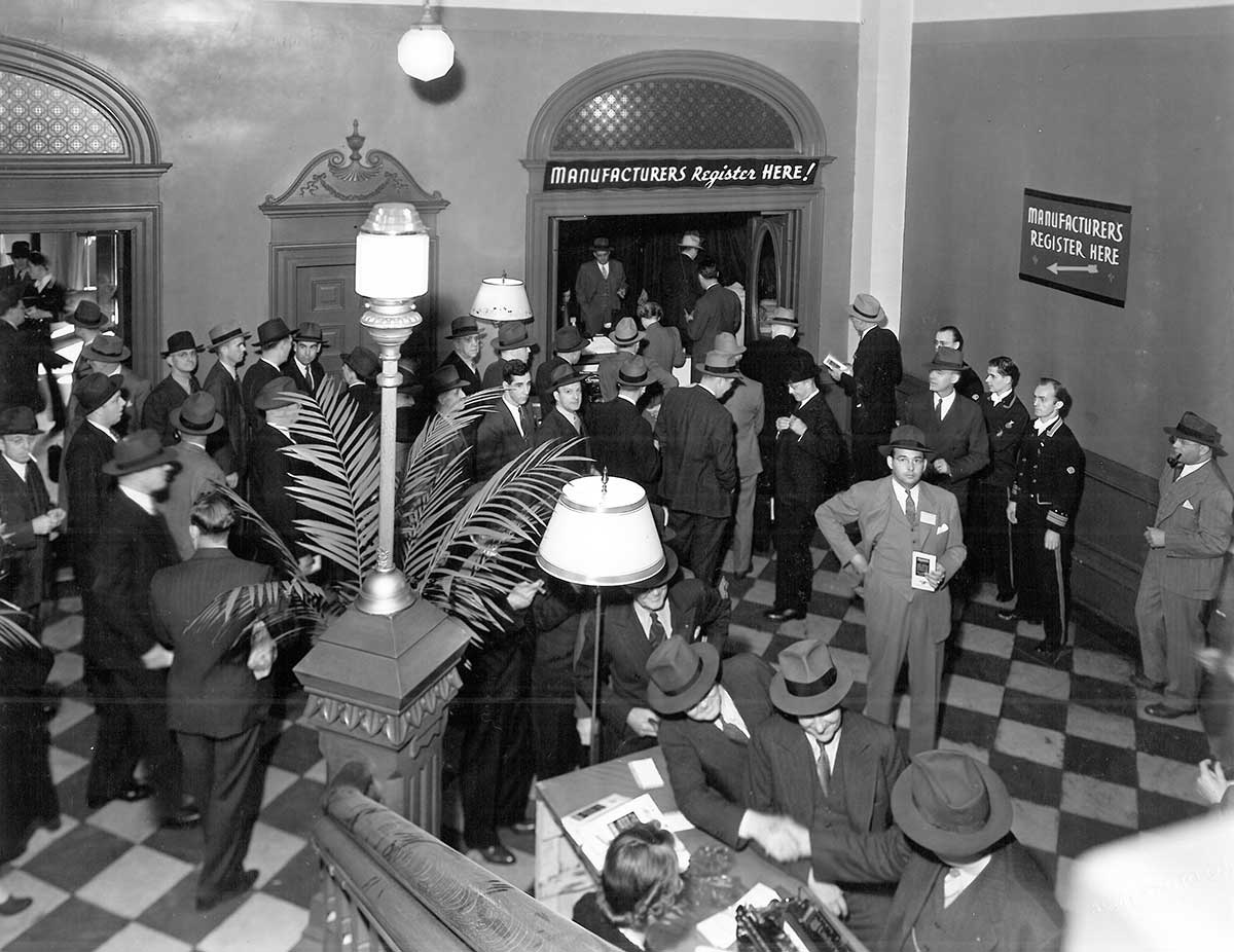 Historic photo showing the north corridor lobby with checkerboard stone at the foot of grand staircase, 1928; large group of men milling around.